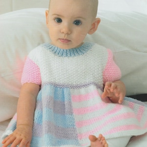 May include: A baby wearing a white, pink, and blue knitted dress with a checkered pattern. The dress has short sleeves and a round neckline.