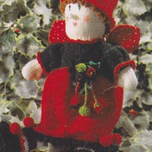 Può includere: Bambola natalizia lavorata a maglia a mano con cappello, vestito e ali rossi. La bambola ha un viso bianco con guance rosate e un maglione nero. La bambola è decorata con accenti verdi e rossi, su uno sfondo di agrifoglio.