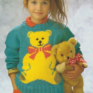 May include: A teal blue knitted sweater with a yellow bear design and a red bow. The sweater is being worn by a young girl who is holding a brown teddy bear.