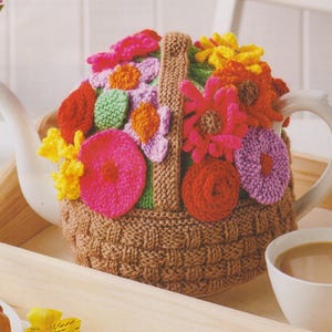 Könnte beinhalten: Eine braune gestrickte Teekannenhülle mit einem Korbgeflechtmuster und bunten gehäkelten Blumen in den Farben Rosa, Gelb, Rot und Grün.