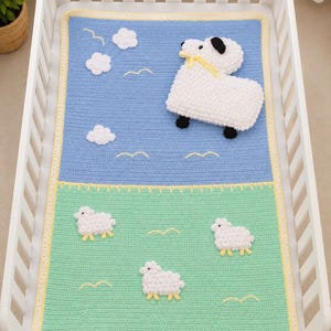 May include: A handmade crochet baby blanket in a crib. The blanket features a blue sky with white clouds and a large sheep applique. The bottom half is green with three sheep appliques. Yellow trim borders the blanket.