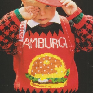 Könnte beinhalten: Ein Kind trägt einen roten Pullover mit einem großen Burger-Design und dem Wort "AMBURG" in Weiß. Der Pullover hat schwarz-rot gemusterte Ärmel, grüne und schwarze Verzierungen und einen weißen Kragen. Das Kind trägt auch eine rote Kappe.