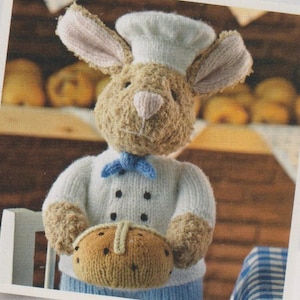 Puede incluir: Un conejo tejido con gorro y uniforme de chef, sosteniendo un pan. El conejo tiene pelaje marrón, orejas rosas y una pajarita azul. El pan es marrón con manchas negras y una raya de color beige claro.