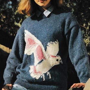 May include: A blue knit sweater featuring a white and pink dove design. The dove is in flight, with its wings spread across the front of the sweater. The sweater has a crew neck and long sleeves. The person is wearing a collared shirt underneath.