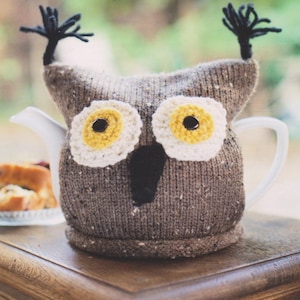 May include: A brown knitted owl teapot cozy with yellow and black eyes. The cozy is on a wooden surface.