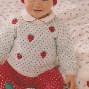 Peut inclure: Un bébé portant un pull blanc et un pantalon rouge et vert à motif fraise. Le bébé porte également un bonnet rouge en tricot avec une fraise verte sur le dessus.