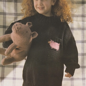Pode incluir: Um suéter preto de malha com um desenho de porco rosa no bolso. O suéter é usado com calças listradas e um brinquedo de porco de malha. A criança tem cabelo ruivo encaracolado e uma faixa.
