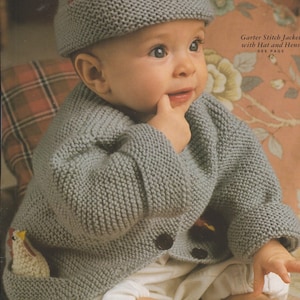 Op de afbeelding: Een baby draagt een grijze gebreide trui met een kip-applicatie op de zak en een bijpassende grijze gebreide muts met een kip-applicatie op de bovenkant. De trui heeft een knoopsluiting. De baby zit op een stoffen met een bloemenpatroon.