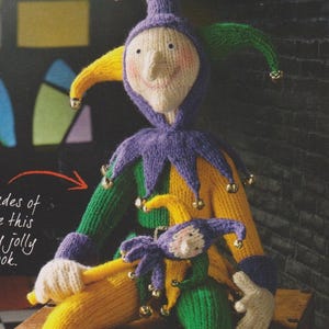 May include: Two knitted jester dolls, one large and one small, are sitting on a wooden surface. The larger jester is wearing a purple, yellow, and green costume with bells. The smaller jester is wearing a purple, green, and yellow costume with bells. Both jesters have a long, pointed nose and a wide smile.