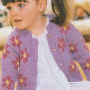 May include: A young girl wearing a purple cardigan with pink and yellow flower embroidery. The cardigan has a button closure and is knitted.