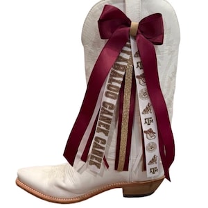 May include: A white cowboy boot adorned with maroon and white ribbons. The ribbons feature text and logos, including "CABALOO CANEK CANEK" and "ATM". A gold glitter ribbon and a maroon bow complete the festive design.