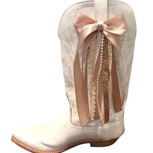 May include: A white cowboy boot with a decorative bow and ribbon embellishment. The bow is a light pink color, and the ribbons include lace and pearl accents. The boot has a pointed toe and a brown sole.