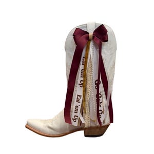 Op de afbeelding: Een witte cowboylaars versierd met een bordeauxrode strik en linten. De linten tonen de tekst "Eat 'em Up" en "Go Cats Go". De laars heeft een puntige neus en een houten hak, met decoratieve stiksels.