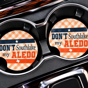 May include: Two round car cup holder coasters featuring an orange and white gingham pattern. The text reads DON'T Southlake my ALEDO in dark blue and orange lettering, accented by a small white star and a white silhouette of the state of Texas.