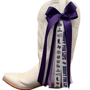 May include: A white cowboy boot with a purple bow and decorative ribbons. The ribbons have the text "HORNY FOR LIFE" and various graphics. The boot has a pointed toe and decorative stitching.