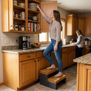 May include: A two-step wooden stool with a dark gray base and natural wood treads, positioned in a kitchen. The stool features a sturdy construction with visible joinery, providing elevation to reach items stored in high wooden kitchen cabinets.