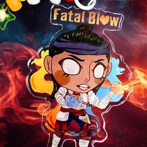 May include: A clear acrylic keychain featuring a cartoon character with brown hair, a blue headband, and a red and white outfit. The character is holding a blue and white orb in one hand and a red and orange orb in the other. The keychain has the text "Fatal Blow" in gold letters with a heart symbol.