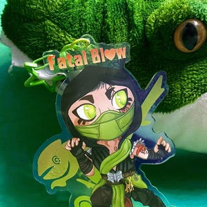 May include: A green and black anime-style character keychain with the text "Fatal Blow" in red and a heart. The character is wearing a green mask and has green eyes. The keychain is on a green background.