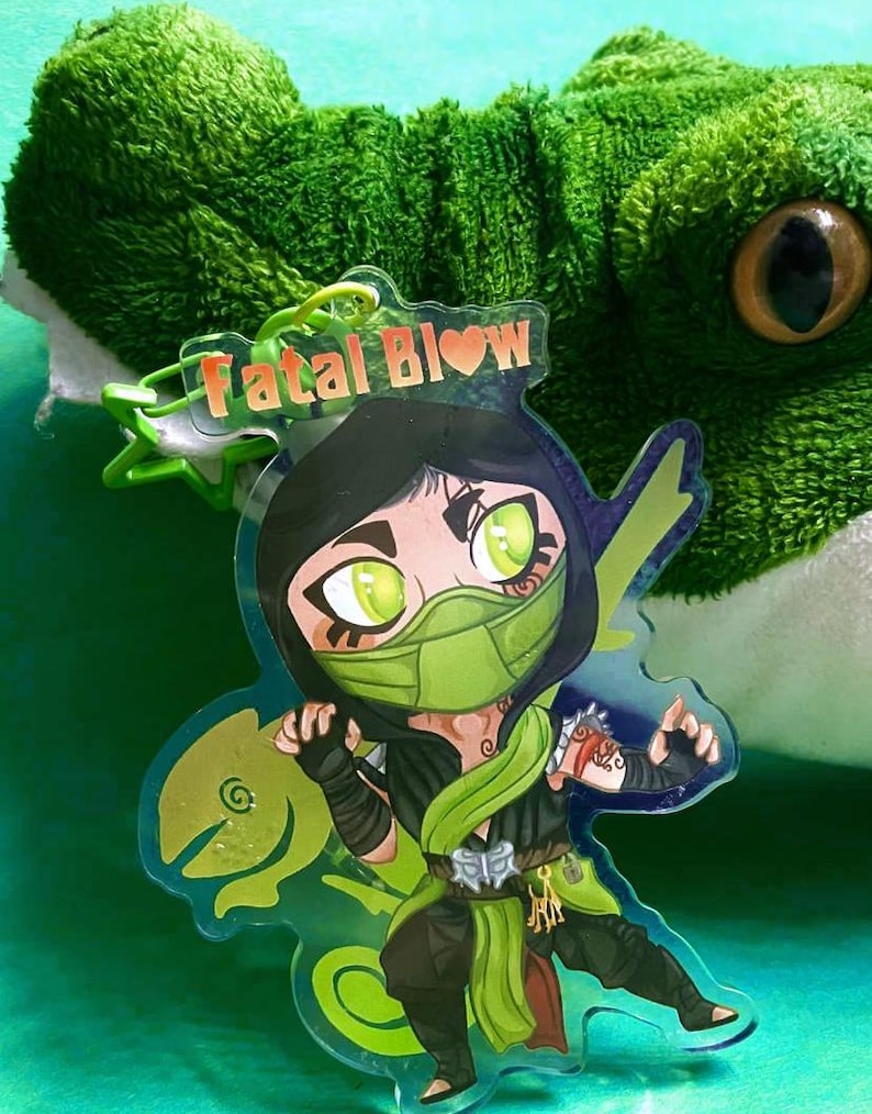 May include: A green and black anime-style character keychain with the text "Fatal Blow" in red and a heart. The character is wearing a green mask and has green eyes. The keychain is on a green background.
