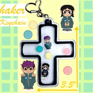 Puede incluir: Un llavero agitador en forma de cruz con un cierre en forma de corazón. El llavero presenta personajes de dibujos animados y acentos coloridos. El texto "Shaker Keychain" se muestra en amarillo. El llavero mide 8,89 cm de largo.