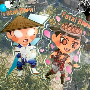 May include: Two anime-style character charms with the text "Fatal Blow" on each. The charm on the left features a character with a white and blue outfit, a brown hat, and a white and blue background. The charm on the right features a character with a red and black outfit, a brown hat, and a pink and white background.