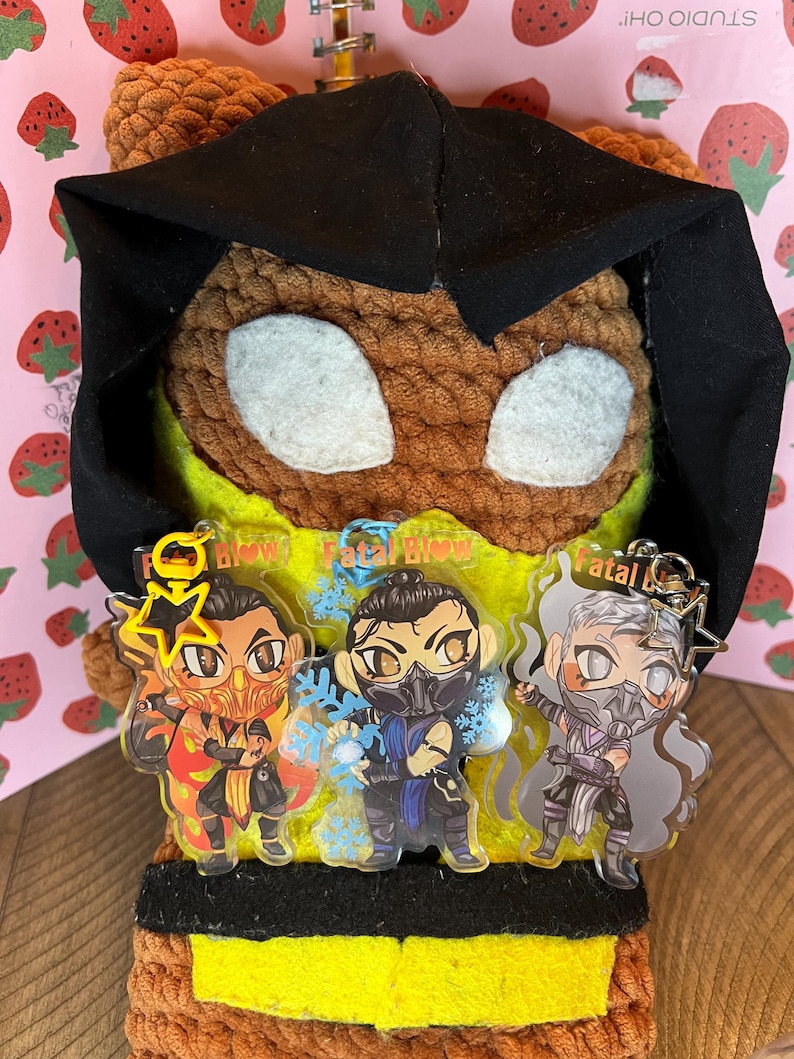 May include: A brown crocheted plush toy wearing a black hooded jacket. The toy is holding three clear acrylic charms with images of characters from the video game Mortal Kombat. The charms are labeled "Fatal Blow", "Fatal Blow", and "Fatal Blow".
