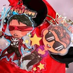 Op de afbeelding: Twee kleurrijke acrylcharms met cartoonfiguren met de tekst "Fatal Blow" op elke charm. De personages dragen een zonnebril en een rood ooglapje.