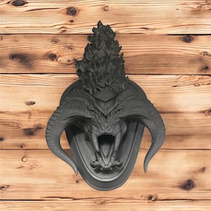 May include: A dark gray, three-dimensional wall sculpture of a demonic creature's head with large horns and an open mouth. The sculpture is mounted on a gray oval plaque, with a textured flame-like element above the head.