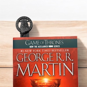 Puede incluir: Un libro negro con el título "Game of Thrones" y el nombre del autor "George R.R. Martin" en la portada. El libro es un best-seller del New York Times y está basado en la serie de HBO. Un marcapáginas negro con una mano sosteniendo una corona está encima del libro.
