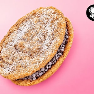 May include: A round oatmeal cookie sandwich filled with dark chocolate chips, featuring a light dusting of white powdered sugar on the top surface. The cookie has a textured, golden-brown appearance and is set against a solid pink background.