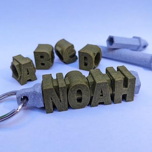 Könnte beinhalten: Ein personalisierter Schlüsselanhänger mit dem Namen "NOAH" in olivgrünen, 3D-gedruckten Buchstaben. Die Buchstaben sind an einer grauen Basis mit einem silbernen Schlüsselring befestigt. Zusätzliche Buchstaben und ein graues Werkzeug sind ebenfalls sichtbar.