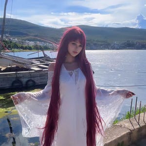 May include: A person with long, deep red hair and bangs, wearing a white dress with lace sleeves, stands near a body of water. The dress has a V-neck and off-the-shoulder sleeves. The background includes a lake, mountains, and a blue sky.