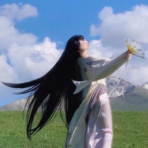 May include: A woman with long black hair wearing a white and pink traditional Chinese dress, holding a white and gold fan, standing in a field with green grass and a blue sky with white clouds.