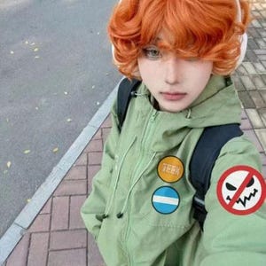 Puede incluir: Una persona con una chaqueta verde claro con parches, una mochila negra y cabello naranja. La chaqueta tiene cremallera y capucha. Los parches incluyen la palabra "GEEK" y un diseño circular con rayas azules y blancas. La persona lleva auriculares blancos.