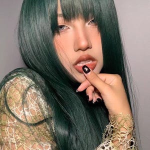 May include: A person with long, dark green hair styled with blunt bangs. They are wearing a sheer, light brown, lacy top.