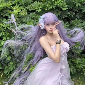 Könnte beinhalten: Eine Person mit einer lavendelfarbenen Perücke mit langem, fließendem Haar, Elfenohren und einem trägerlosen, hellvioletten Kleid. Die Person ist mit floralen Haaraccessoires und einem Armband geschmückt. Der Hintergrund zeigt üppiges grünes Laub.