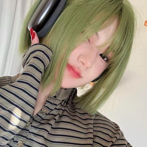 Puede incluir: Una persona con cabello verde usando auriculares negros y una camisa a rayas. Los auriculares son negros con una almohadilla gris. La camisa es beige con rayas azul oscuro.