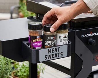 May include: A black plastic side shelf attachment for a Blackstone griddle featuring a rectangular storage bin labeled with the text MEAT SWEATS in bold black letters. The bin holds three spice seasoning jars including Grill Mates and Weber brands.