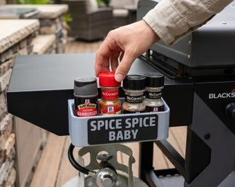 May include: A light gray rectangular spice rack attached to a black outdoor griddle. The rack features a black label with the white text SPICE SPICE BABY and holds four McCormick spice bottles. The rack is made of a matte plastic material.