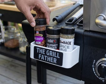May include: A white rectangular magnetic spice rack attached to a black grill, featuring the text THE GRILL FATHER in bold black letters. The rack holds three spice containers, including McCormick Mesquite, Weber Gourmet Burger, and Rib Rack Spice Rub.