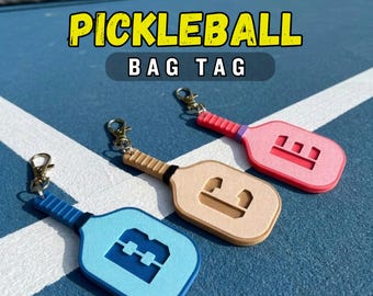 Puede incluir: Tres etiquetas para bolsos con forma de pala de pickleball en azul claro, beige y rosa, cada una con un cierre de mosquetón de metal y un diseño recortado en la cara de la pala. El texto PICKLEBALL BAG TAG aparece en amarillo y blanco.