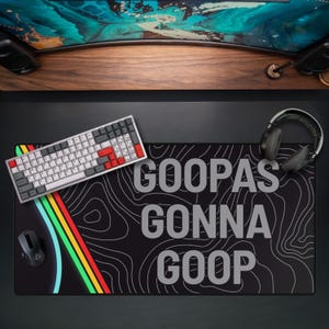 May include: Black mousepad with the text "GOOPAS GONNA GOOP" in gray. A keyboard with red and gray keys, a mouse, and headphones are on the pad. The pad has a topographic design and colorful stripes.