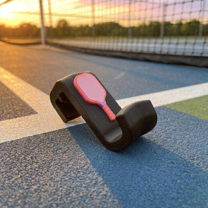 May include: A black pickleball paddle holder with a pink paddle design. The holder is on a blue court with a net in the background. The paddle is pink with a red handle and a purple accent.