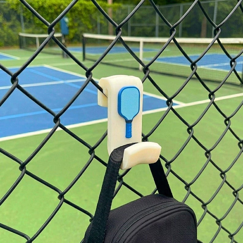 May include: A white and blue pickleball paddle-shaped hook attached to a black chain-link fence. A black bag hangs from the hook. The background shows a pickleball court with a green surface and blue lines.
