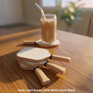 May include: A set of three pickleball paddles with white faces, latte brown bodies, and black bands on the handles. The paddles are stacked together, and a glass of iced coffee with a straw sits on a separate paddle in the background.