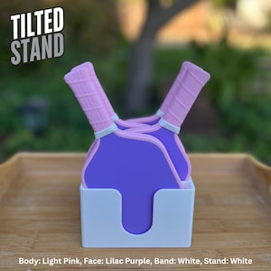 May include: A white stand holding two purple pickleball paddles with light pink handles. The paddles feature a grid pattern on the handles and a white band separating the handle from the paddle face. The text TILTED STAND appears in the upper left.