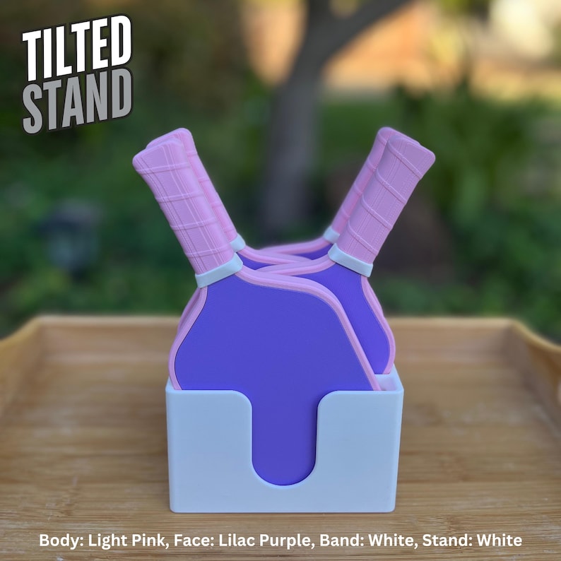 May include: A white stand holding two purple pickleball paddles with light pink handles. The paddles feature a grid pattern on the handles and a white band separating the handle from the paddle face. The text TILTED STAND appears in the upper left.