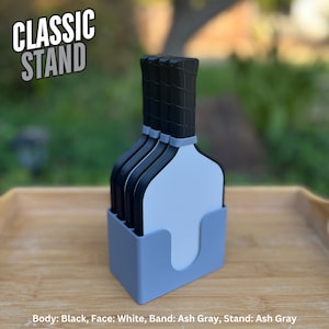 May include: A set of four pickleball paddles with black bodies, white faces, and ash gray bands, held in an ash gray stand. The text CLASSIC STAND is visible in the upper left corner. Text at the bottom reads: Body: Black, Face: White, Band: Ash Gray, Stand: Ash Gray.