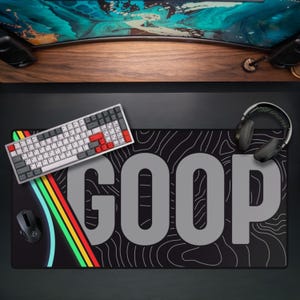 May include: Black GOOP mousepad with a white and gray keyboard, a black mouse, and black headphones. The mousepad has a topographic design and the word GOOP in large gray letters. A rainbow stripe design is on the left side.