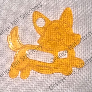 May include: A yellow plastic cutout of a cartoon dog with a happy expression. The dog is running and has a long tail. The cutout is designed for use in cross-stitch projects.